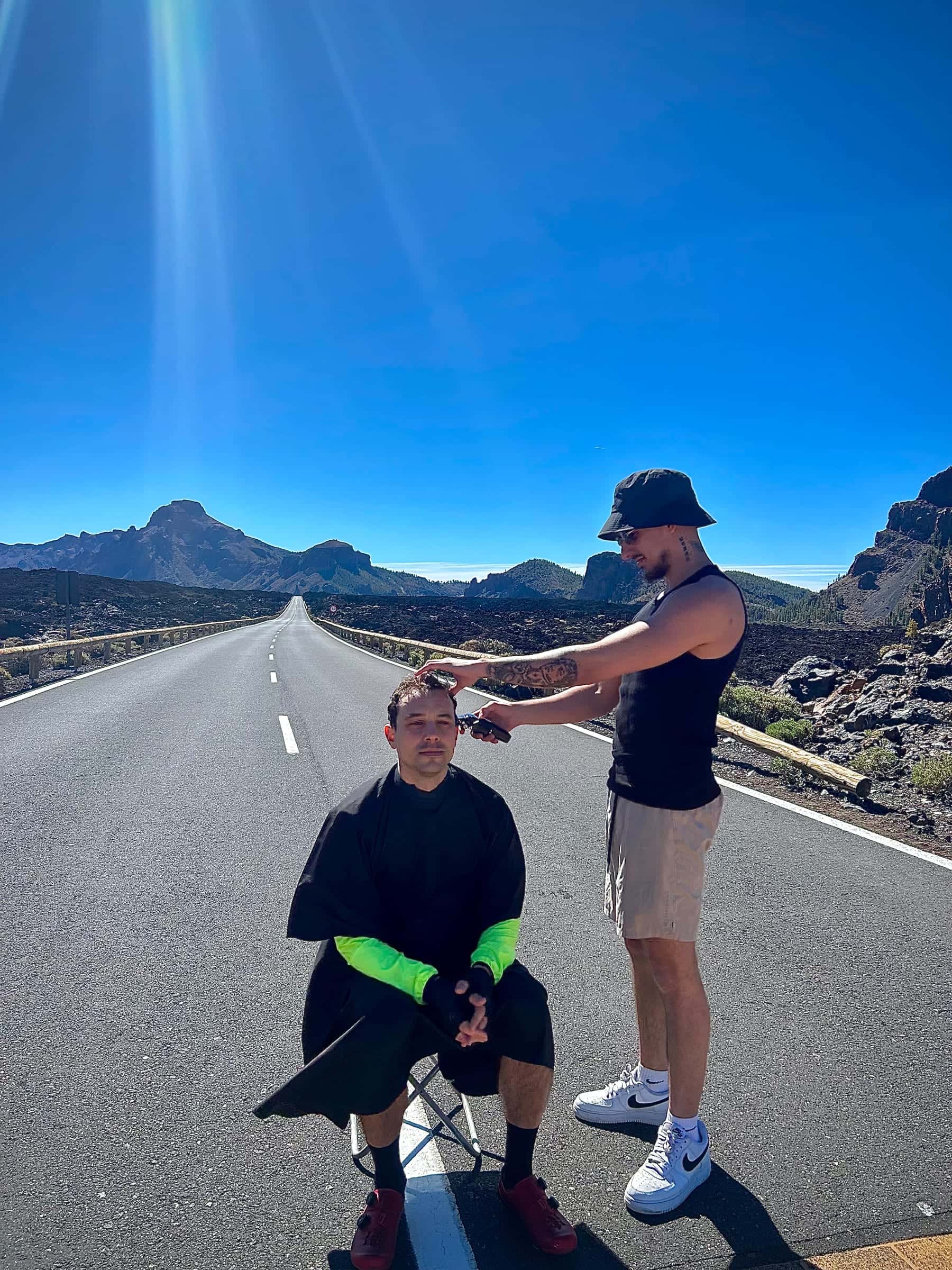 Krystian cutting a seated client's hair in the middle of an empty mountain road on Tenerife, the volcanic peaks around Mount Teide and a deep blue sky behind them.