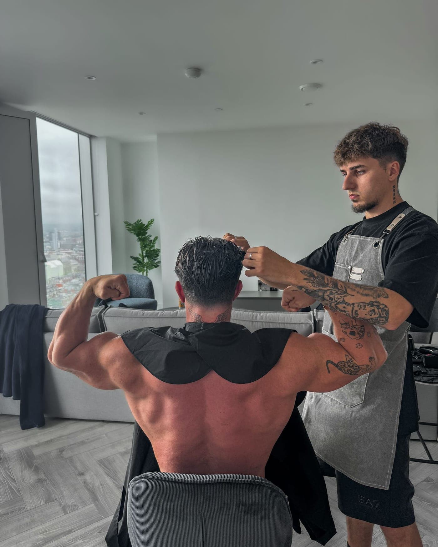 Krystian cutting a client's hair in a high-rise flat, with the Manchester skyline through the window behind them.
